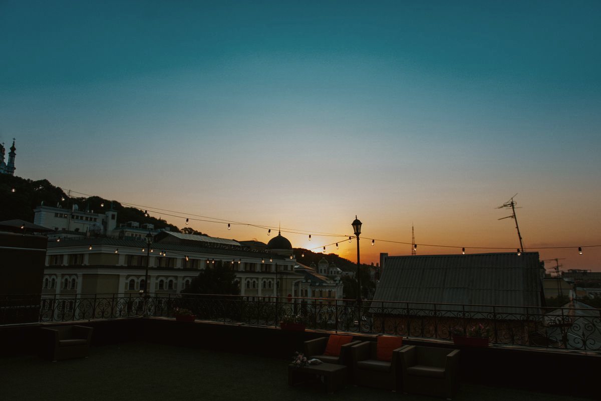 A rooftop lounge with low seating at sunset, rooftops and string lights beyond